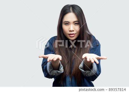 Portrait of serious beautiful brunette asian young woman in casual blue denim jacket with makeup standing raised arms and looking at camera. indoor studio shot, isolated on light grey background. 89357814