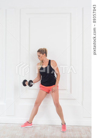 Young adult motivation athletic beautiful bodybuilder woman in pink shorts and black top standing, holding dumbells doing exercise for arms at the gym, lean on white wall. indoor, studio shot Young adult motivation athletic beautiful bodybuilder woman in pink shorts and black top standing, holding dumbells doing exercise for arms at the gym, lean on white wall. indoor, studio shot 89357843