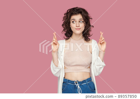 Portrait of funny hopeful beautiful brunette young woman with curly hairstyle in casual style standing with crossed finger and looking away, hope to win. indoor studio shot isolated on pink background 89357850