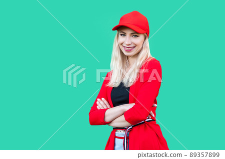 Portrait of happy beautiful blond young hipster style woman in red blouse and cap, standing, crossed arms and looking at camera with toothy smile. indoor studio shot, isolated on green background. 89357899