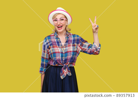 Portrait of happy modern stylish mature woman in casual style with hat, eyeglasses standing with victory or peace sign, looking at camera with toothy smile. studio shot isolated on yellow background. 89357913