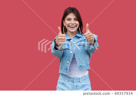 Portrait of happy satisfied beautiful brunette young woman with makeup in denim casual style standing, thumbs up and looking at camera with toothy smile. indoor studio shot, isolated on red background Portrait of happy satisfied beautiful brunette young woman with makeup in denim casual style standing, thumbs up and looking at camera with toothy smile. indoor studio shot, isolated on red background 89357934