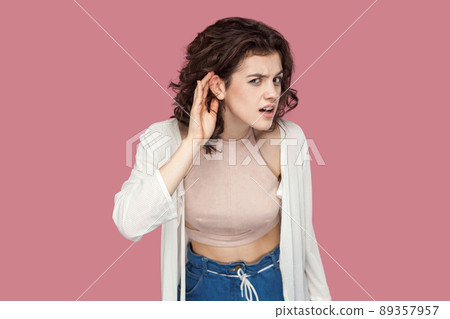 what? tell me louder. Portrait of attentive brunette young woman with curly hairstyle in casual style standing with hand gesture on ear and looking at camera. studio shot isolated on pink background. 89357957