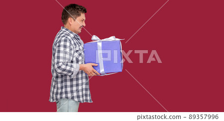 Side view portrait of happy modern middle aged man in white t-shirt and in checkered shirt standing and giving you big heavy gift box with toothy smile. indoor studio shot on isolated red background. Side view portrait of happy modern middle aged man in white t-shirt and in checkered shirt standing and giving you big heavy gift box with toothy smile. indoor studio shot on isolated red background. 89357996