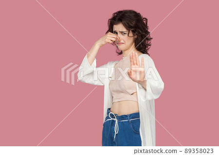 Portrait of unhappy brunette young woman with curly hairstyle in casual style standing pinching her nose and showing stop gesture and looking at camera. indoor studio shot isolated on pink background. Portrait of unhappy brunette young woman with curly hairstyle in casual style standing pinching her nose and showing stop gesture and looking at camera. indoor studio shot isolated on pink background. 89358023