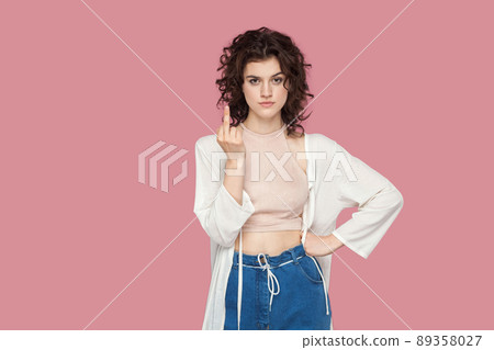 Portrait of careless brunette young woman in curly hairstyle, casual style standing with middle finger fuck sing and looking at camera with poker face. indoor studio shot isolated on pink background. Portrait of careless brunette young woman in curly hairstyle, casual style standing with middle finger fuck sing and looking at camera with poker face. indoor studio shot isolated on pink background. 89358027