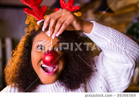 Joyful brazil charming woman in toy deer horns holding red ball gift box while at cozy room 89366569