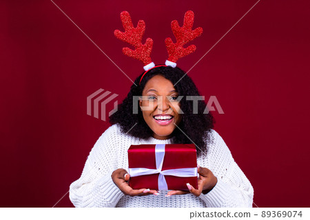 Joyful brazil charming woman in toy deer horns holding red ball gift box while at cozy room 89369074