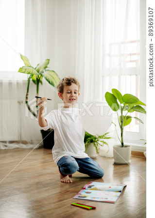 A cute boy draws a picture on a piece of paper sitting on the floor in a room. Childhood, leisure, free time A cute boy draws a picture on a piece of paper sitting on the floor in a room. Childhood, leisure, free time 89369107