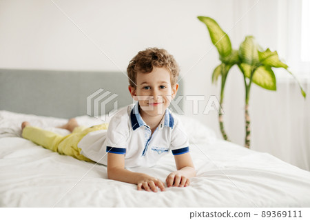 The cute boy is lying on his stomach on the bed on a white blanket, looking at the camera, smiling The cute boy is lying on his stomach on the bed on a white blanket, looking at the camera, smiling 89369111