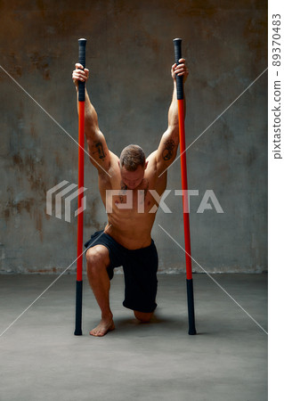 A fighter shirtless in black shorts, stands in a fighting artificially pose and holds a fight stick on studio background. A fighter shirtless in black shorts, stands in a fighting artificially pose and holds a fight stick on studio background. 89370483