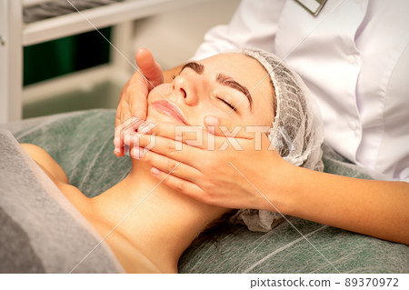 Beautiful caucasian young woman receiving a facial massage with closed eyes in spa salon, close up. Relaxing treatment concept Beautiful caucasian young woman receiving a facial massage with closed eyes in spa salon, close up. Relaxing treatment concept 89370972
