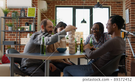 Diverse group of friends having fun with game activity, playing board games with wooden building tower on table. Men and women enjoying play with square blocks and pieces for entertainment. 89372452