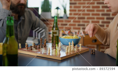 Cheerful people having fun with chess play at board games gathering together, drinking beer and eating snacks. Group of friends enjoying intelligence match for entertainment at home. Cheerful people having fun with chess play at board games gathering together, drinking beer and eating snacks. Group of friends enjoying intelligence match for entertainment at home. 89372594
