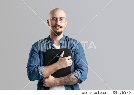 Confident handsome balded beard with musctache man wearing casual clothes holding clipboard isolated over gray background. Confident handsome balded beard with musctache man wearing casual clothes holding clipboard isolated over gray background. 89376580