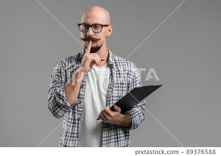 Pensive handsome balded beard with musctache man wearing casual clothes holding clipboard isolated over gray background.  89376588
