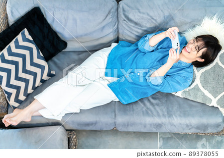 A woman with glasses lying on the sofa and watching a smartphone video A woman with glasses lying on the sofa and watching a smartphone video 89378055