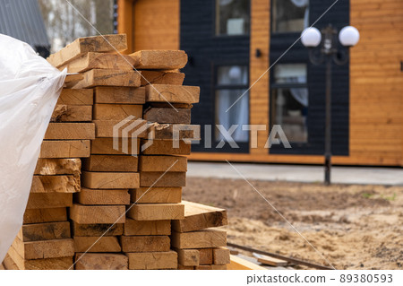 Construction site. In the foreground lies the building material of the board. In the background is a wooden house built from such boards Construction site. In the foreground lies the building material of the board. In the background is a wooden house built from such boards 89380593