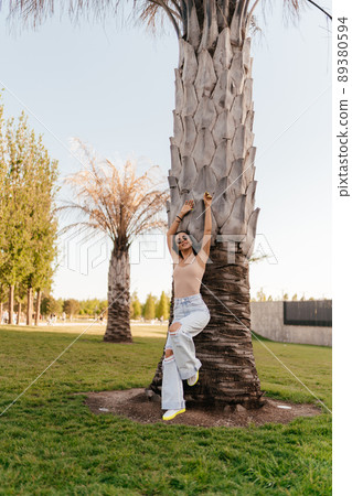 A beautiful young girl in glasses, a beige T-shirt and white jeans stands leaning on a palm tree in the park. The student stands with her arms up and her right leg bent. Summer walks in the park. The A beautiful young girl in glasses, a beige T-shirt and white jeans stands leaning on a palm tree in the park. The student stands with her arms up and her right leg bent. Summer walks in the park. The 89380594