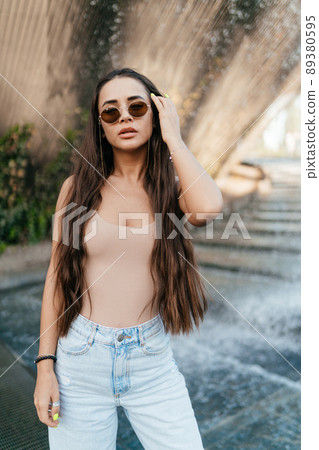 A beautiful girl with dark long hair in a beige T-shirt and blue jeans, with glasses standing against the background of a fountain in the park. The girl corrects her hair with her left hand. Walks in A beautiful girl with dark long hair in a beige T-shirt and blue jeans, with glasses standing against the background of a fountain in the park. The girl corrects her hair with her left hand. Walks in 89380595