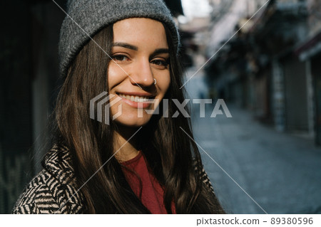 A girl with dark straight long hair in a gray coat, hat and burgundy T-shirt. Portrait of a happy student close-up against the background of a gray street. Travel to Istanbul during the pandemic. The A girl with dark straight long hair in a gray coat, hat and burgundy T-shirt. Portrait of a happy student close-up against the background of a gray street. Travel to Istanbul during the pandemic. The 89380596
