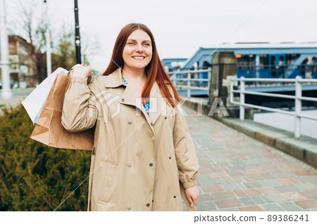 Happy young redhead woman holding shopping paper bags in hands, walking at city street, black friday. Urban lifestyle concept. Delivery carrying. 89386241