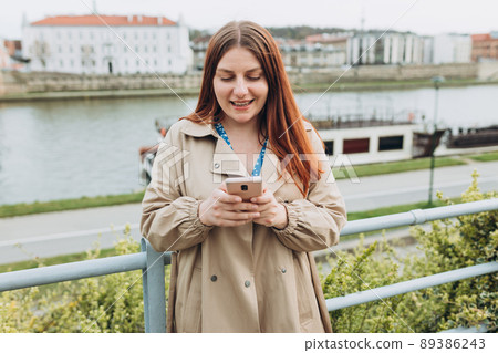 Beautiful happy redhead woman is using a smart phone and smiling outdoors. Urban lifestyle concept. Girl walking on city street checks her smartphone. Spring time Beautiful happy redhead woman is using a smart phone and smiling outdoors. Urban lifestyle concept. Girl walking on city street checks her smartphone. Spring time 89386243