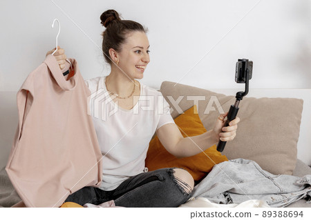 Young woman sitting on the bed at home and taking photo of her old clothes to sell them online. Selling on website, e-commerce. Reuse, second-hand concept. Conscious consumer, sustainable lifestyle. Young woman sitting on the bed at home and taking photo of her old clothes to sell them online. Selling on website, e-commerce. Reuse, second-hand concept. Conscious consumer, sustainable lifestyle. 89388694