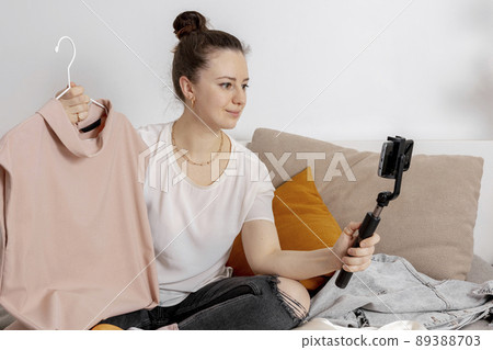 Young woman sitting on the bed at home and taking photo of her old clothes to sell them online. Selling on website, e-commerce. Reuse, second-hand concept. Conscious consumer, sustainable lifestyle. 89388703