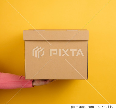 female hand holding closed brown cardboard box on yellow background, delivery 89389119