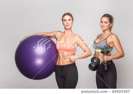 Two activity strong woman looking at camera and posing with gym equipment. Studio shot, isolated on light gray background Two activity strong woman looking at camera and posing with gym equipment. Studio shot, isolated on light gray background 89405497