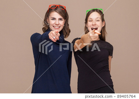 Women standing near each other and pointing fingers at camera. Studio shot, light brown background Women standing near each other and pointing fingers at camera. Studio shot, light brown background 89405764