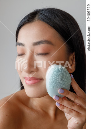 Smiling Asian woman using face massager in studio Smiling Asian woman using face massager in studio 89406729