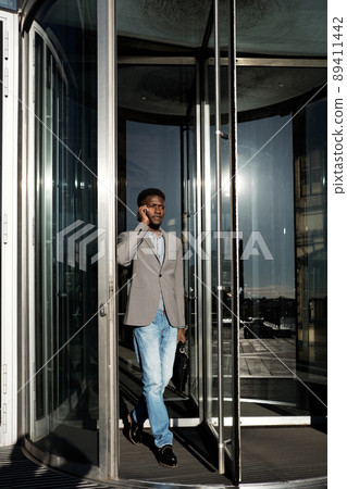 End of working day. African American manager going through glass revolving door and talking on smartphone End of working day. African American manager going through glass revolving door and talking on smartphone 89411442