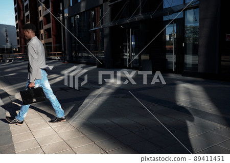 End of working day. Side view of African American manager with case moving away from modern business center, shadows seen on pavement 89411451