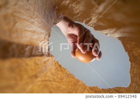 A hand takes a chicken egg out of a paper bag. A mature man's hand holds a brown egg over an open brown bag. Shot from the bottom up. Close up. Selective focus 89412348