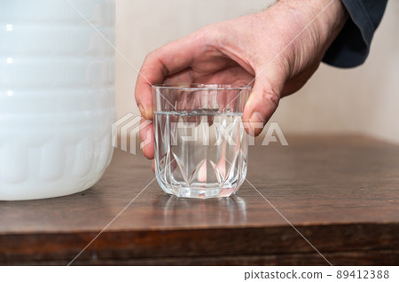 A man holds a glass of drinking water in his hand. A large white plastic bottle of fresh water is next to him on the table. Inside the room. 89412388