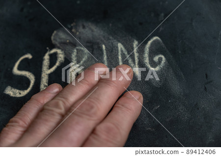 A man's hand wipes off the word SPRING. The word written on a black surface in yellow chalk. Handwritten text. A man's hand wipes off the word SPRING. The word written on a black surface in yellow chalk. Handwritten text. 89412406