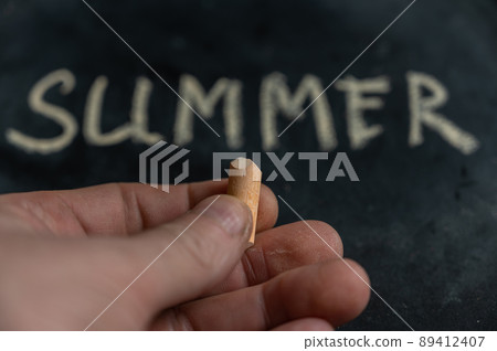 Close-up of a man's hand holding yellow chalk. The word SUMMER written on a black surface in the background. Handwritten text. Close-up of a man's hand holding yellow chalk. The word SUMMER written on a black surface in the background. Handwritten text. 89412407