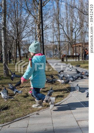 A girl scares the pigeons in the city park. A flock of birds eats sitting on the path. A child runs and scares the birds. 89412410