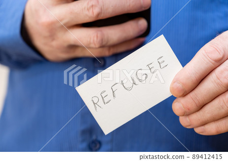 Refugee. Inscription in jagged gray letters on paper. Man holding up two white paper rectangles with handwritten text. Close-up of the hand of a mature man. Refugee. Inscription in jagged gray letters on paper. Man holding up two white paper rectangles with handwritten text. Close-up of the hand of a mature man. 89412415