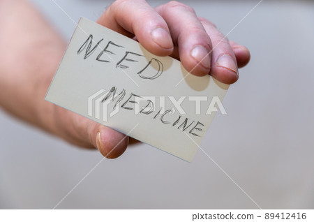 Need medicine. Words written in jagged letters. A man's hand holds a white paper rectangle with text against a gray background Need medicine. Words written in jagged letters. A man's hand holds a white paper rectangle with text against a gray background 89412416
