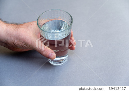 A man's hand holds a tall glass of water against a gray background. A cone-shaped glass of clear drinking water. Selective focus. V A man's hand holds a tall glass of water against a gray background. A cone-shaped glass of clear drinking water. Selective focus. V 89412417