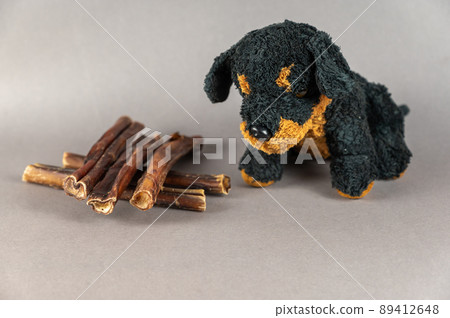 Pet treats and a children's toy opposite the gray background. Natural dried treats and a black stuffed dog. Bully sticks. Pet supplies. 89412648