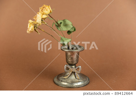 Old candle holder and yellow roses against a brown background. Melchior alloy candle holder. A branch of dried yellow flowers. 89412664