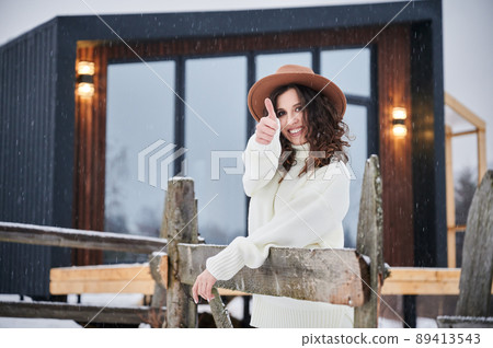 Curly young woman showing thumbs up near modern barn house in the mountains. Happy female tourist enjoying winter holidays in new cottage. Curly young woman showing thumbs up near modern barn house in the mountains. Happy female tourist enjoying winter holidays in new cottage. 89413543