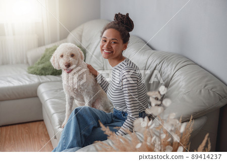 Smiled pretty mixed-race female sitting at cozy room on sofa with white poodle dog Smiled pretty mixed-race female sitting at cozy room on sofa with white poodle dog 89414237