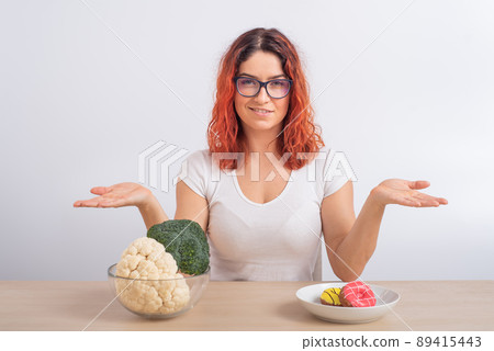 Caucasian woman prefers healthy food. Redhead girl chooses between broccoli and donuts on white background. 89415443
