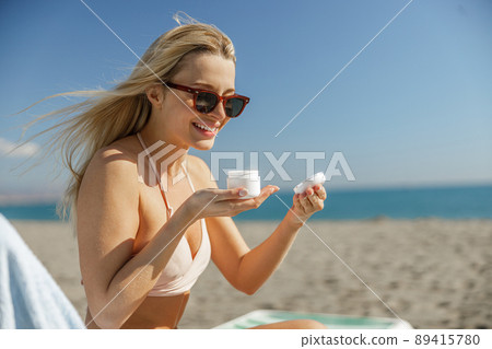 Excited lady in sunglasses opening a jar with body cream while relaxing on beach 89415780