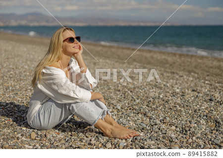 Dreamy woman in casual wear and sunglasses enjoying summertime, sitting barefoot on sea coast 89415882